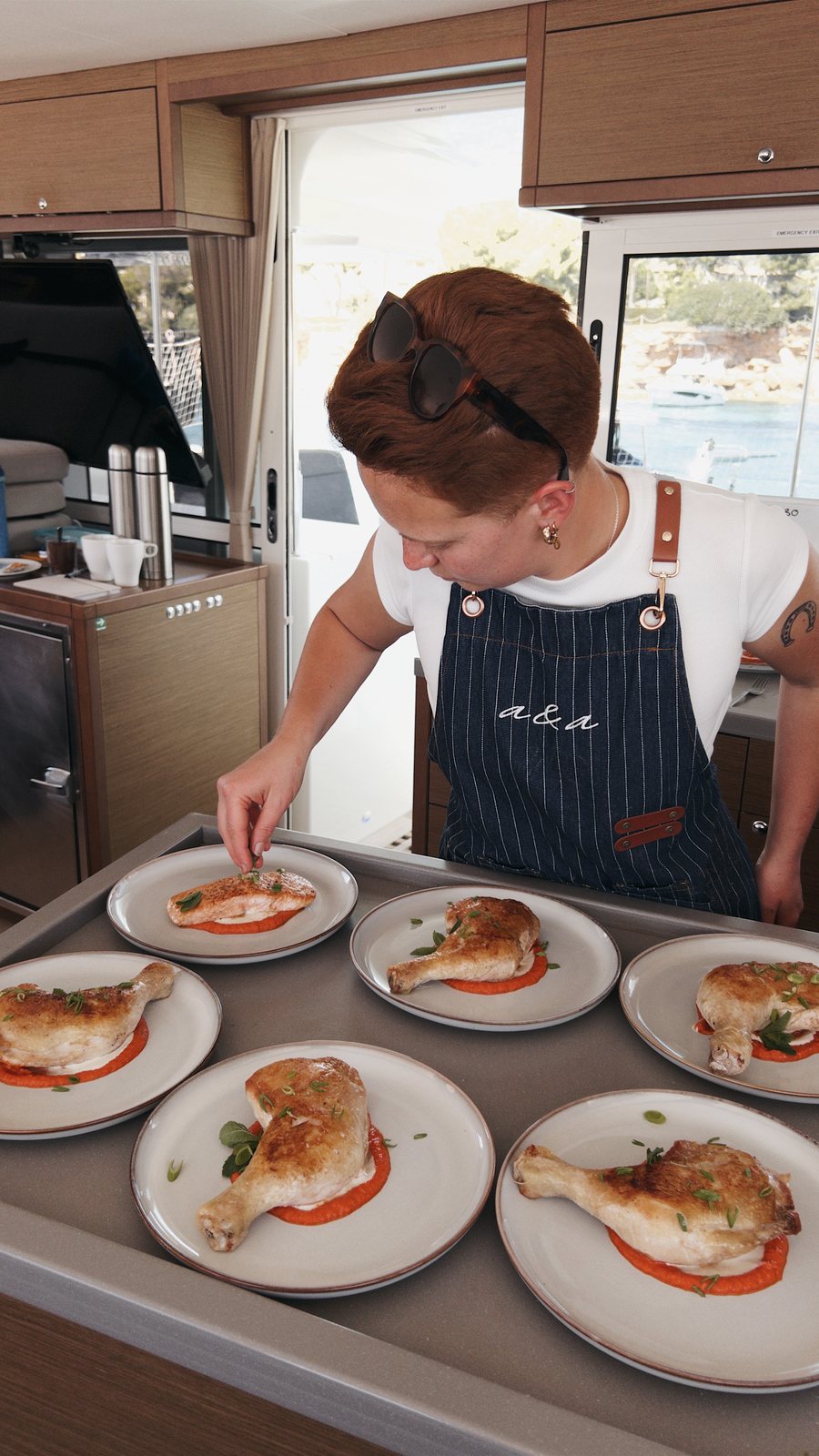 A chef in the school's striped denim apron, sunglasses pushed up on her head, bends over the galley counter of a catamaran finishing six plates of roast chicken on a red sauce; the boat's saloon windows and the water outside sit behind her.
