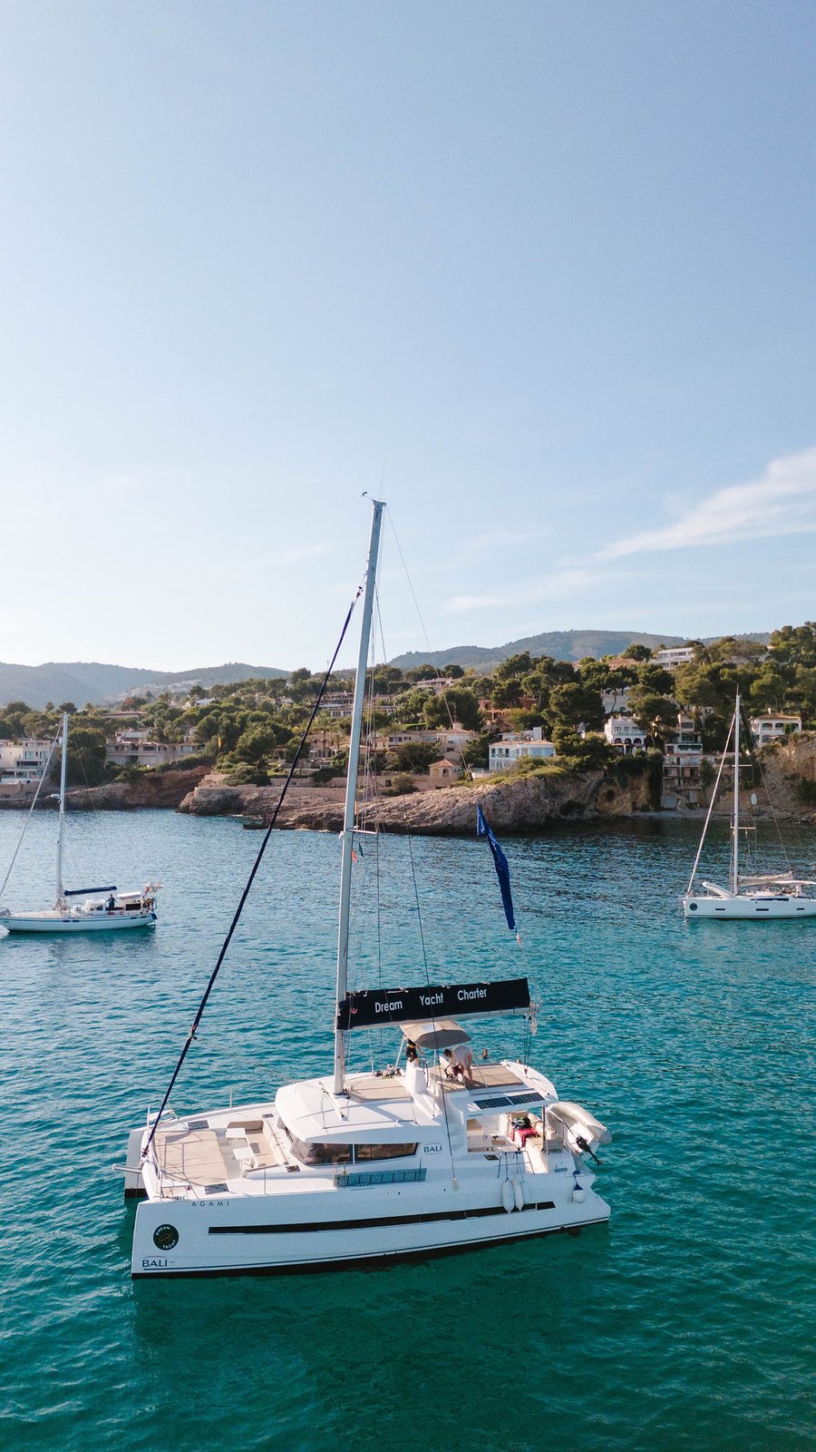 A white sailing catamaran at anchor in a turquoise cove, seen from above, with two more sailing boats moored behind it and a wooded headland lined with houses beyond.
