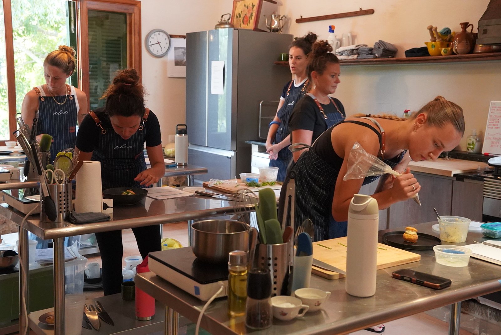 A bright kitchen with two rows of steel benches and portable induction hobs; five women in the school's striped aprons work at them, one piping onto a plate in the foreground, with a tall fridge, a wall clock and a window onto a garden behind.