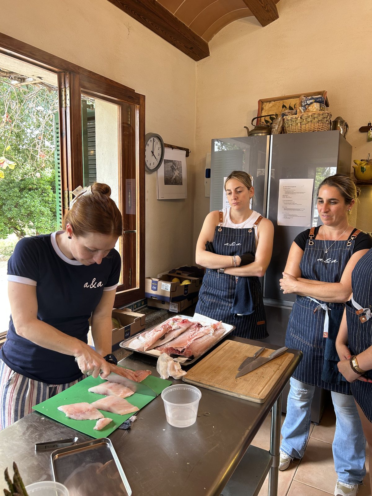 A student in a navy T-shirt and striped apron fillets a white fish on a green board at a steel bench, a tray of whole fish beside her, while two more students in the same aprons stand behind with their arms folded, watching her knife.