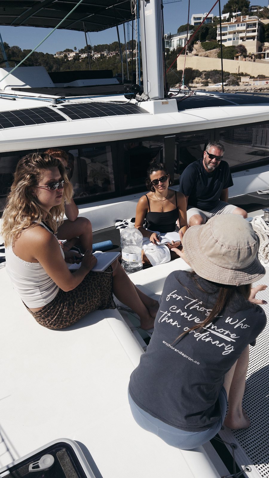 Four people sit in a circle on the foredeck of a catamaran in the sun, notebooks and water bottles between them; one in a sun hat has her back to the camera, the hillside houses of a Mallorca bay behind the boat.