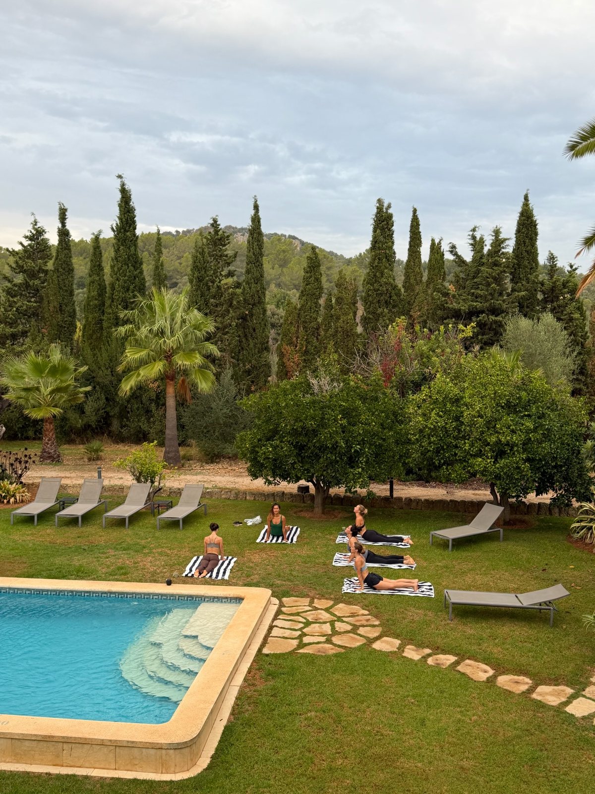 A garden seen from above in early morning: a swimming pool with stone steps, a lawn where a few people stretch on mats, sun loungers along a path, and a line of cypresses and palms against wooded hills.