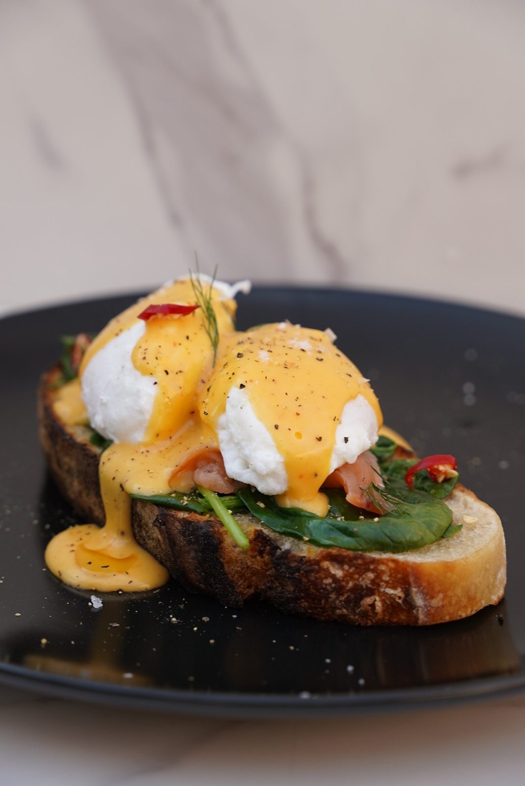 Two poached eggs under hollandaise on toasted sourdough with spinach and smoked salmon, cracked pepper and a sprig of dill, on a black plate.