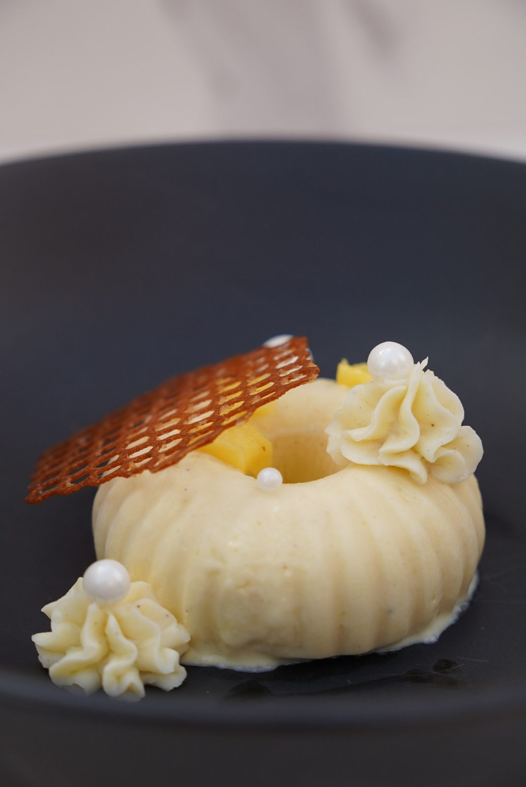 A ridged cream dome on a black plate, with a lace tuile leaning on it, cubes of mango, rosettes of piped cream and small white pearls.