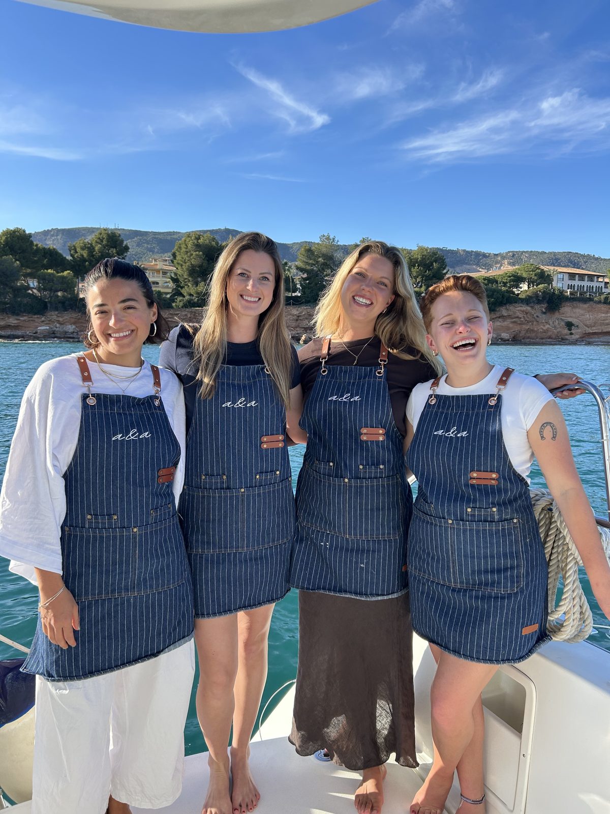 Four women in the school's striped denim aprons stand arm in arm on the stern of a boat, laughing, with a bay and wooded hills behind them.