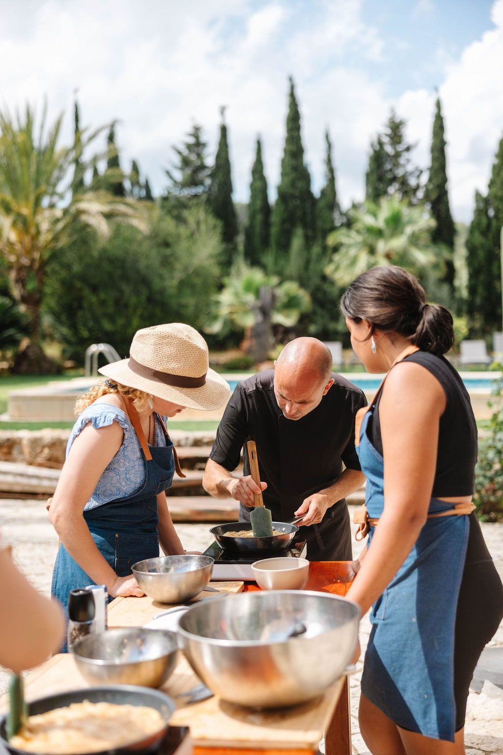 Three people at an outdoor table by a pool: a man in a black T-shirt stirs a pan with a spatula while two women in aprons lean in, steel bowls in front of them and cypresses and palms behind.
