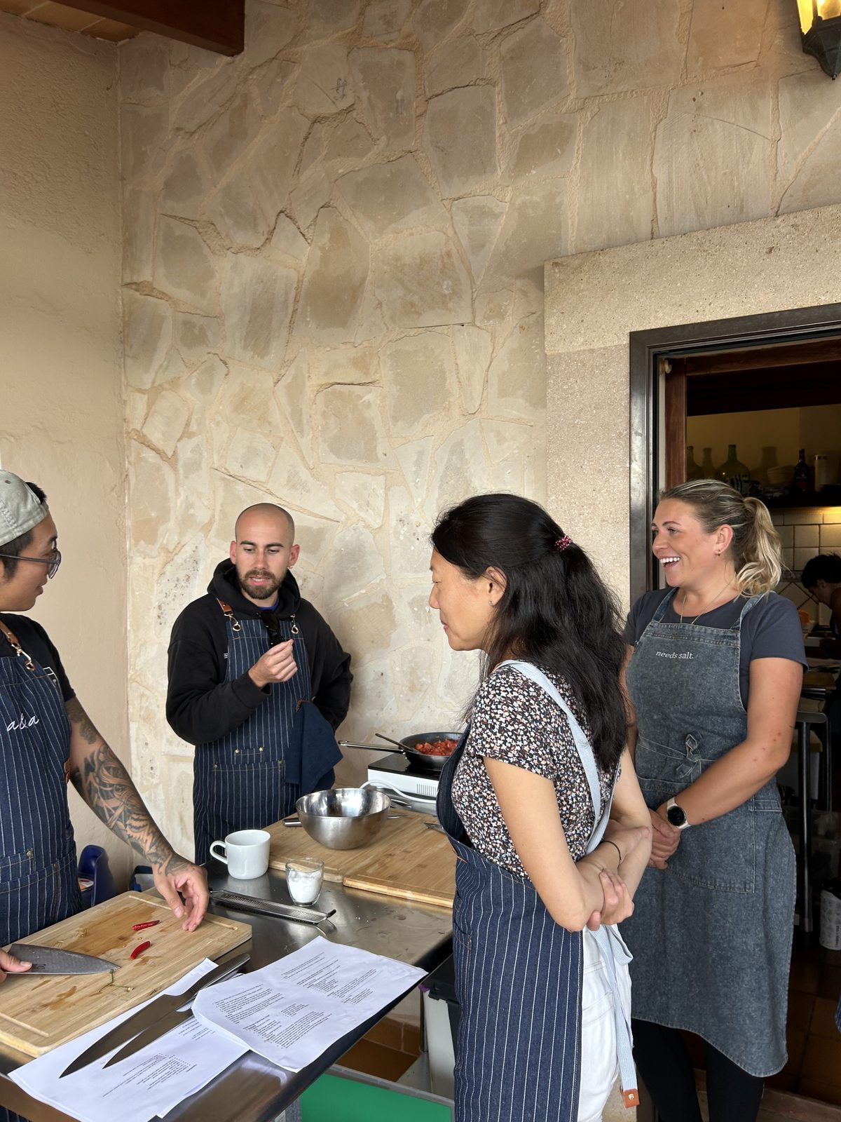 Four people in striped aprons at a steel bench under a stone wall: a man in a black hoodie explains, a woman listens with her hands clasped, another smiles, and a third works a board with recipes and knives laid out.