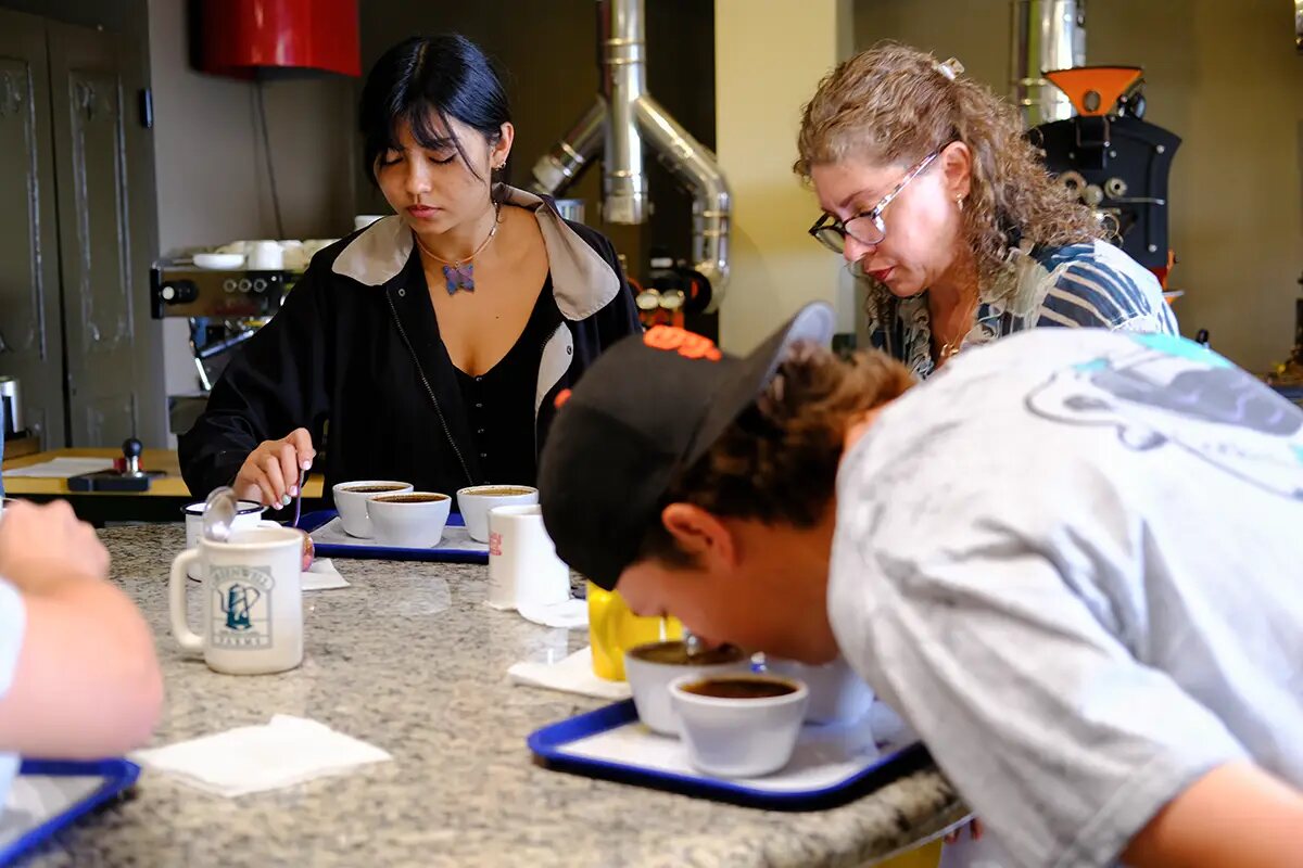 Three students at a granite cupping counter. A young woman spoons from a row of white cupping bowls on a blue tray, a woman in glasses watches beside her, and a young man in a cap bends low over his own bowls in the foreground. A coffee roaster and its steel ducting stand behind them.