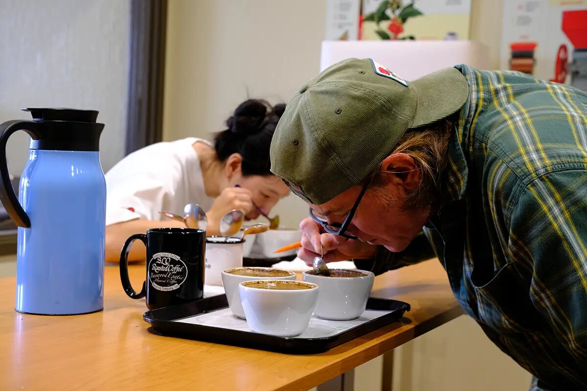 A man in a green cap and glasses leans right down over three white cupping bowls on a black tray, a spoon in his hand. Behind him a woman sips from a spoon over her own bowls.