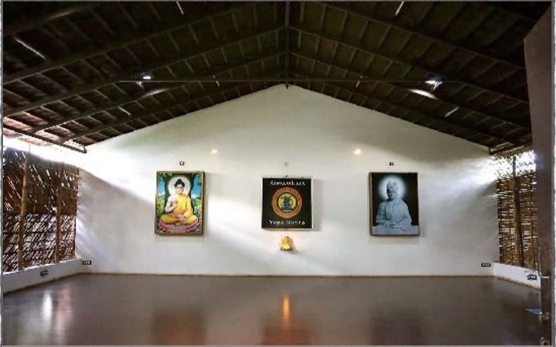 The school's empty practice hall: a high timber roof over a white gable wall hung with three framed pictures — a painted Buddha, the black Atmavikasa Yoga Kutira board and a portrait of Swami Vivekananda — a brass figure on a shelf below the centre board, and a dark polished red floor.