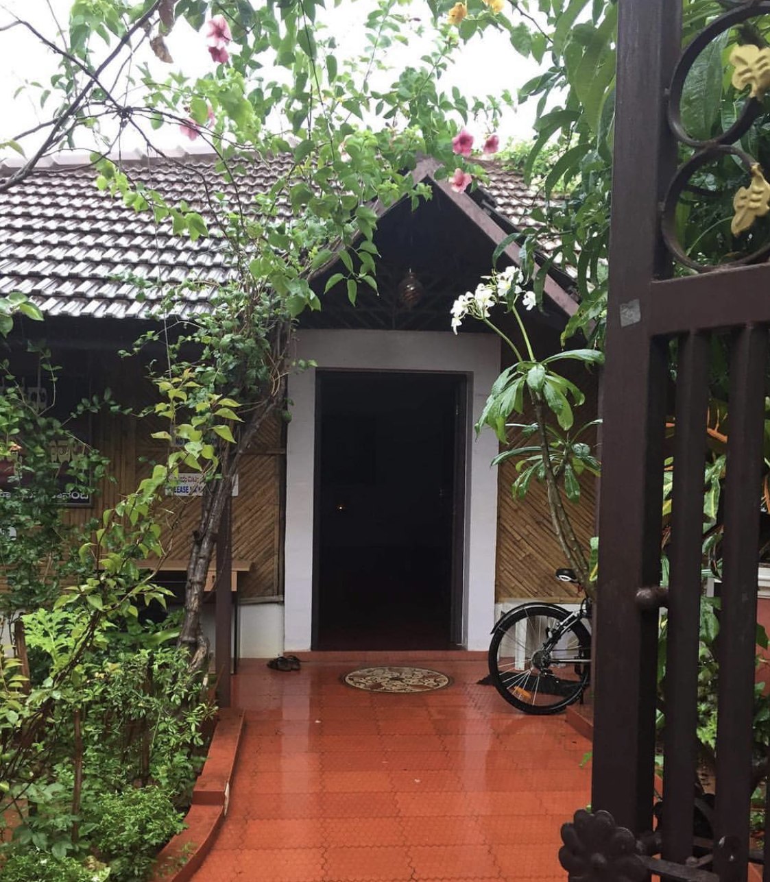 The entrance seen from the garden gate: a tiled roof over bamboo-panelled walls, a white doorway standing open onto darkness, a pair of sandals on the terracotta-tiled path at the threshold, a bicycle against the wall beside the door, hibiscus and frangipani overhead, and a small notice half hidden by leaves.