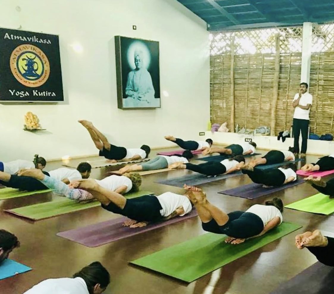 About twenty practitioners lie face down on coloured mats across a dark red floor, all lifting chest, arms and legs clear of the ground at once in the locust pose, below the black Atmavikasa Yoga Kutira board, while a man in a white shirt stands at the far right, hands together, watching.