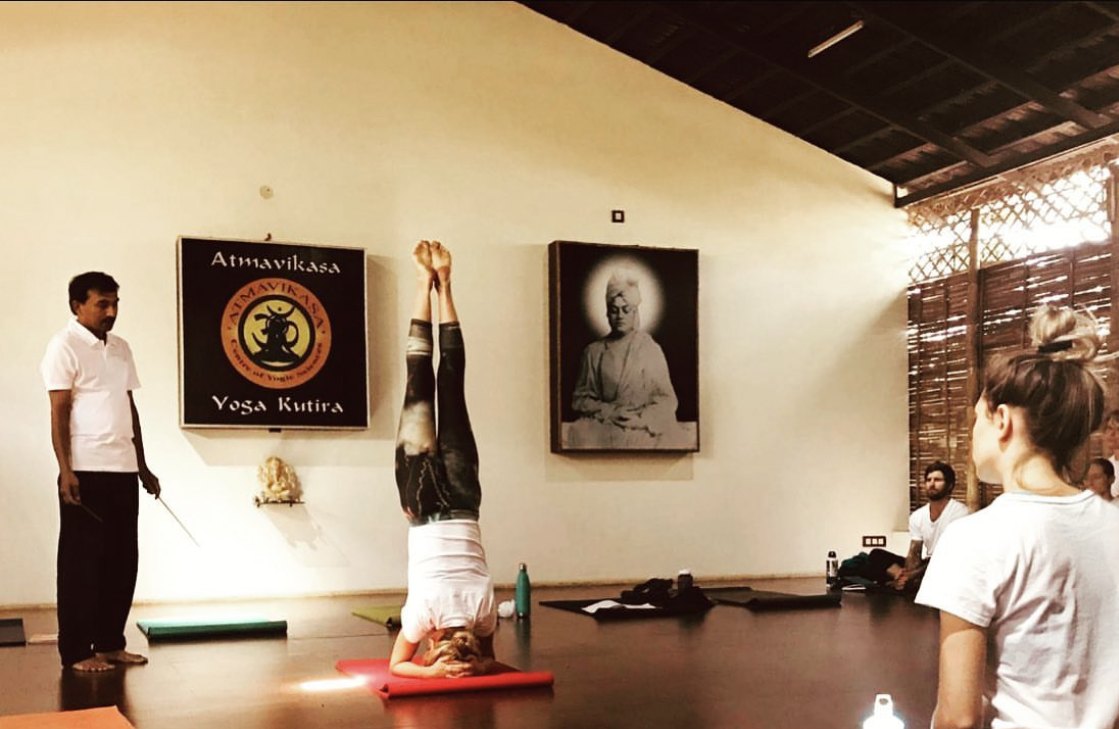 A practitioner holds a headstand on a red mat in the hall, legs straight above them and clasped hands on the floor, while Venkatesha stands at the left with a thin pointer stick held down at his side, watching; two others stand at the right with their backs to the camera.