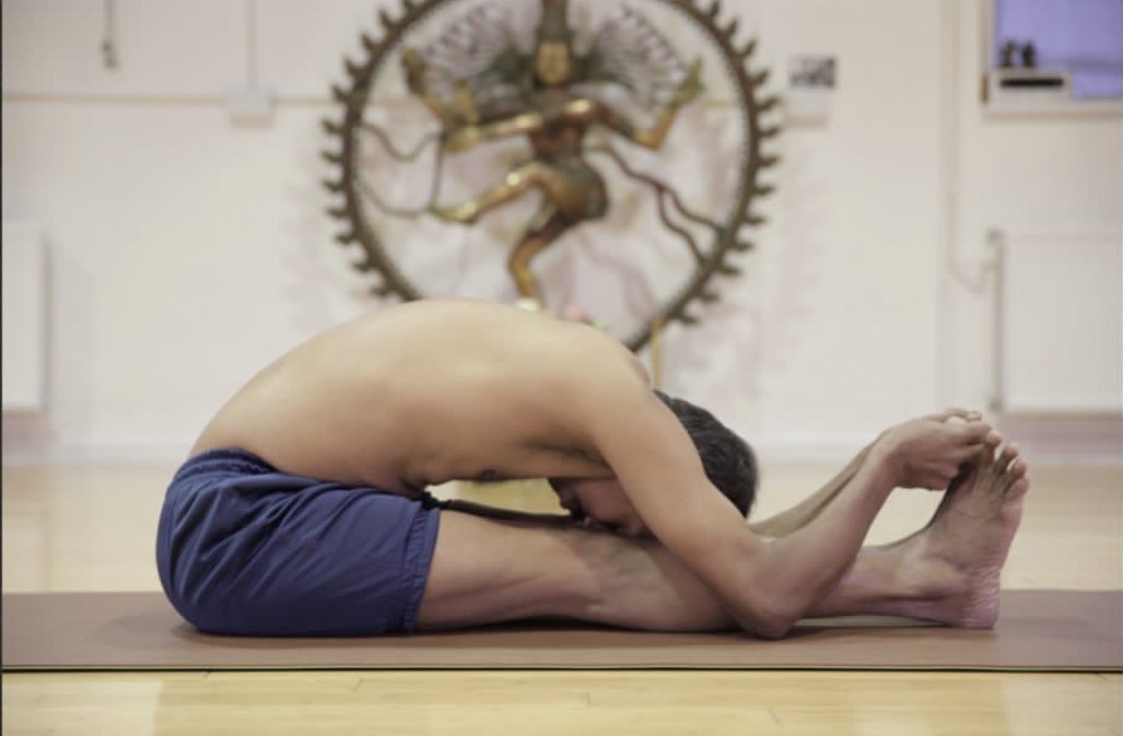 A deep seated forward fold seen from the side at floor level: the practitioner's chest and face lie flat along his straight legs, his back one arc, both arms reaching past his feet, and behind him, out of focus, a bronze Nataraja — Shiva dancing in a ring of flame.