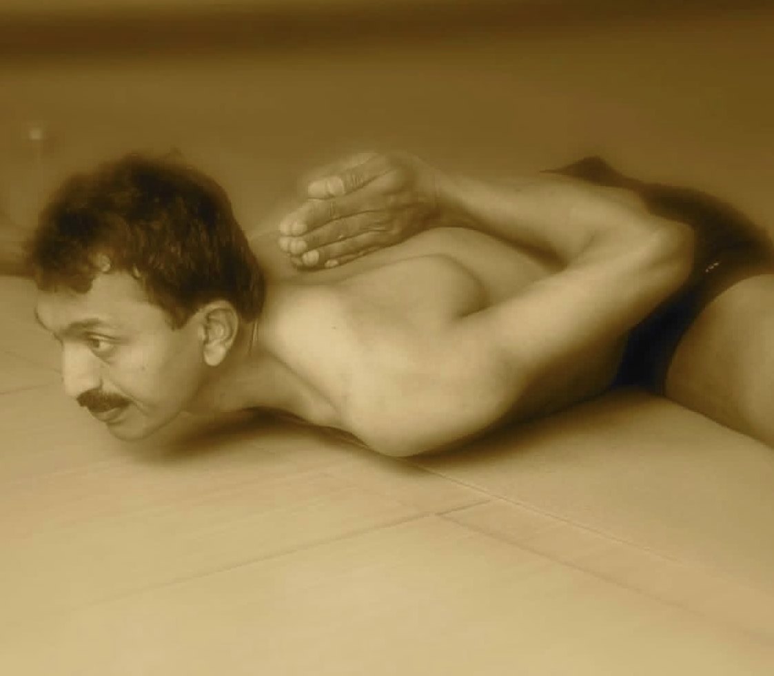 A warm sepia photograph of Yogacharya Venkatesha lying face down on a pale tiled floor, chin and chest to the ground, both arms folded behind his back with the hands drawn up together high between his shoulder blades, his head turned slightly, his eyes open and his expression calm.
