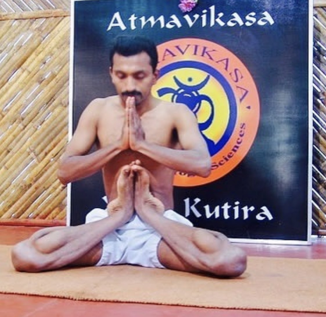 A man in white cotton and no shirt sits with his legs folded and the soles of his feet drawn up and pressed together before his chest, his palms joined above them so that hands and feet mirror each other, against the dark Atmavikasa Yoga Kutira board behind him.
