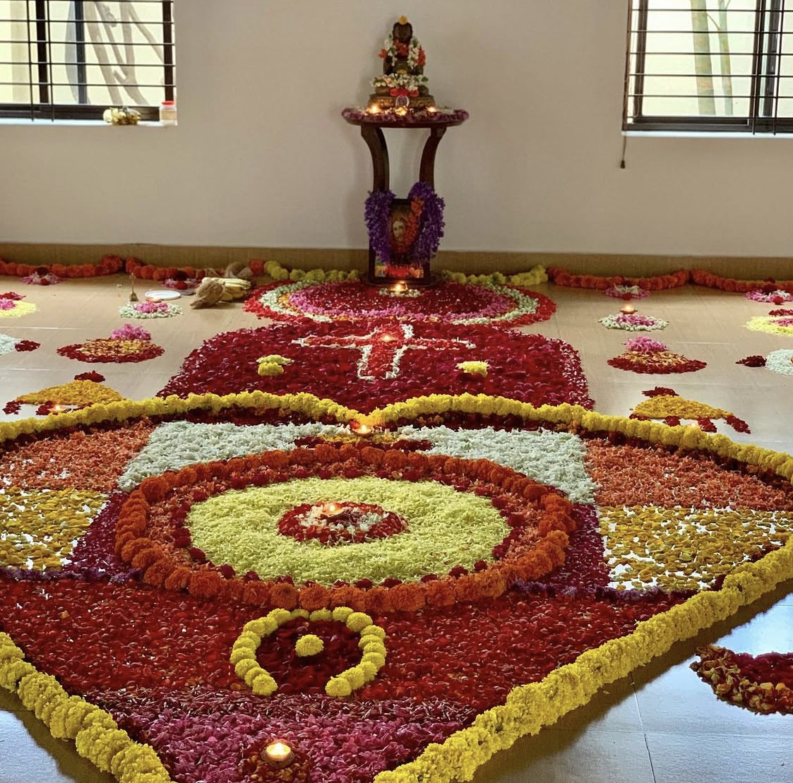 A floor covered end to end by an enormous mandala laid entirely in flower petals, in concentric rings of marigold yellow, deep red, orange, white and pale green, with oil lamps burning in it, and a wooden stand at the far end carrying a garlanded figure above a framed photograph.