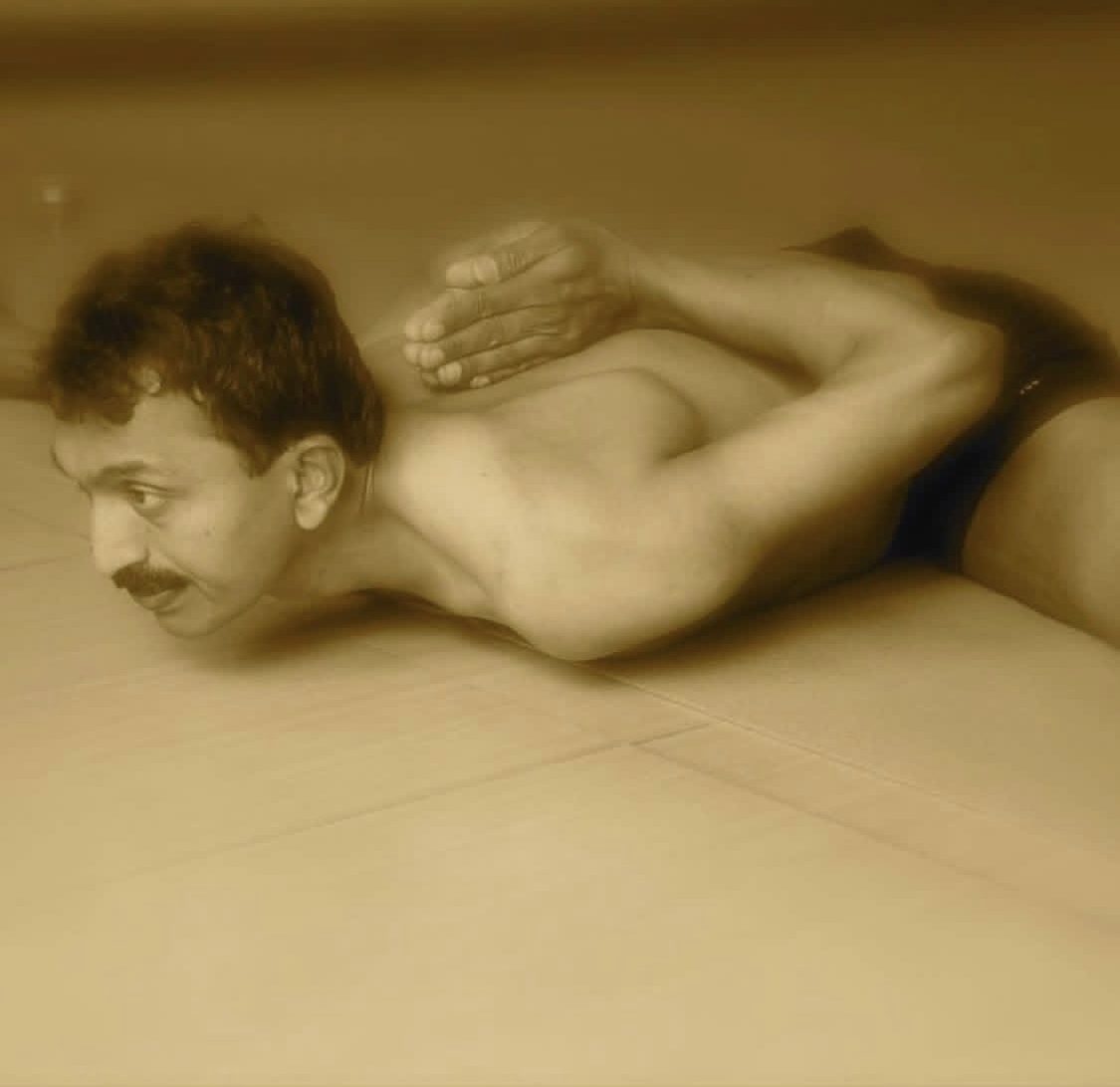 A man lying face down on a pale tiled floor in a bound backbend: the arms are folded up behind the back with the palms pressed together between the shoulder blades, the chest and chin lifted clear of the floor. The photograph is toned sepia.