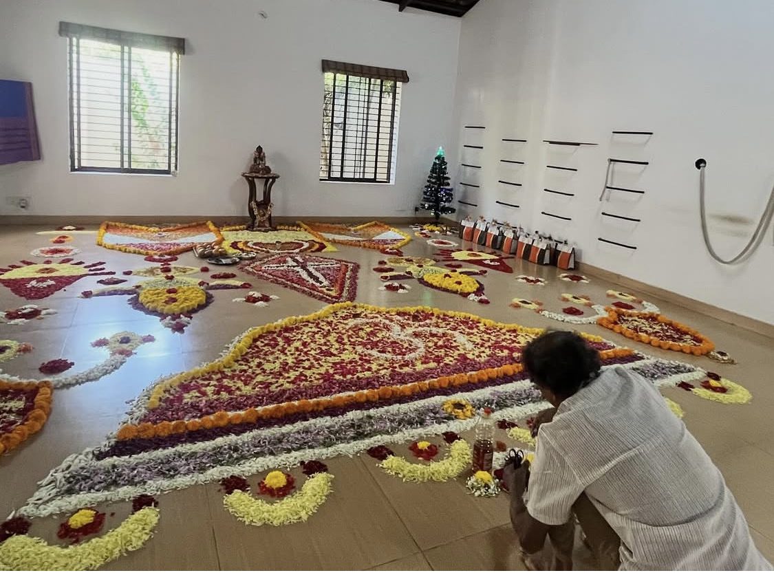 The practice hall with a flower mandala half laid across its floor in concentric bands of marigold, rose, purple and white. A person kneels at the near edge with their back to the camera, setting petals by hand. Behind them a small shrine stands between two shuttered windows, and rope-and-timber props hang on the far wall.