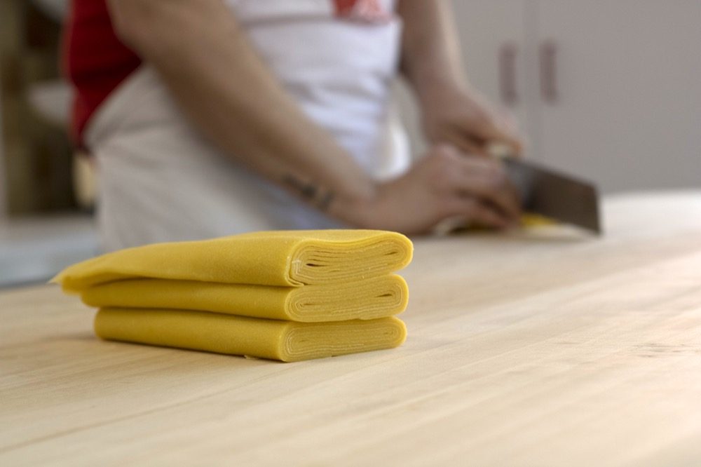 A sheet of yellow egg pasta folded into three flat layers, sharp in the foreground on a pale wooden bench, with a pair of hands and the blade of a long knife out of focus behind it.