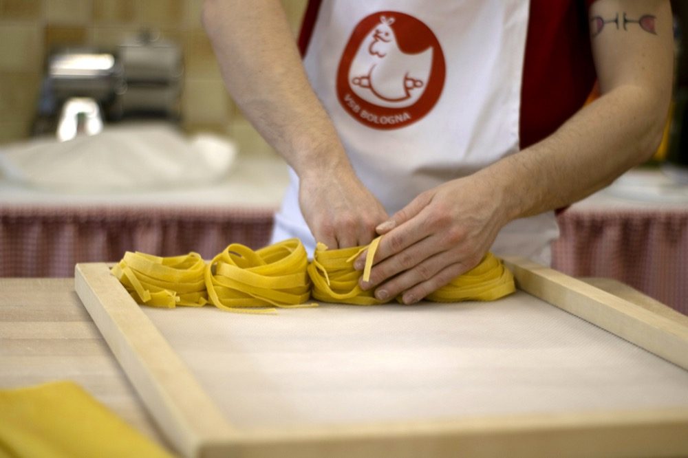 Two hands lifting a fold of freshly cut yellow tagliatelle off a wooden board, the ribbons opening as they rise; the person wears a white apron carrying the school's red roundel, and a hand-crank pasta machine stands out of focus behind.
