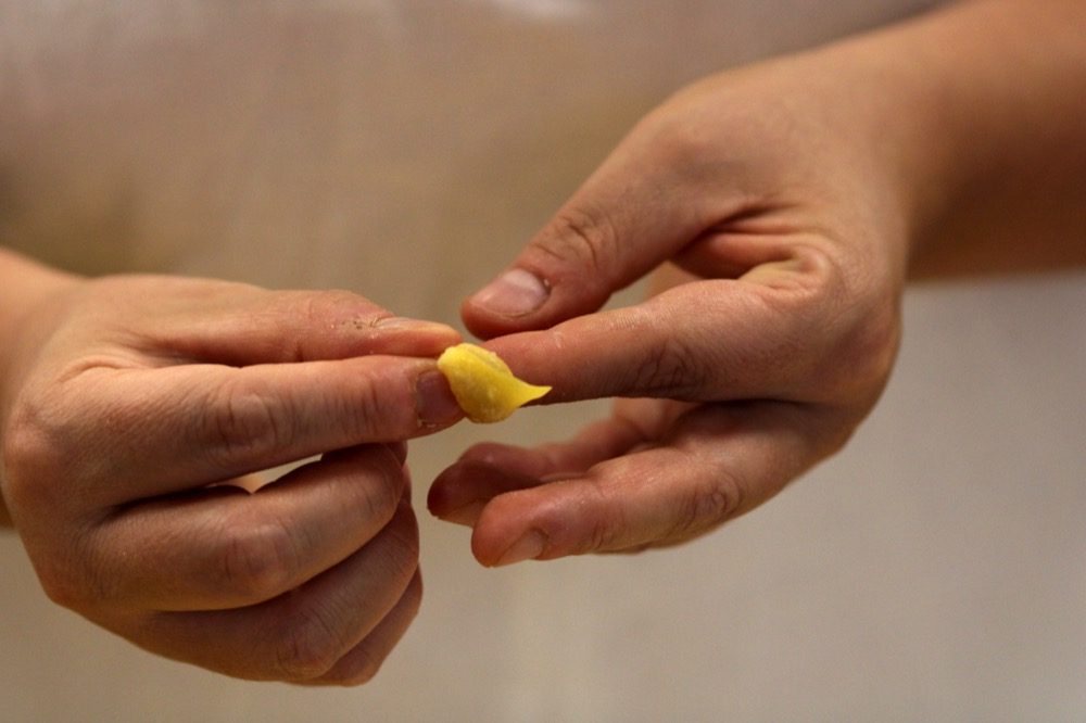 A close view of two hands closing a single small tortellino: the square of yellow pasta is wrapped around the tip of one finger and pinched shut by the other hand, against a plain pale background with nothing else in the frame.