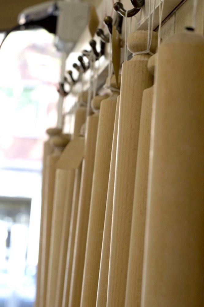A row of long wooden rolling pins standing on end in a line against a bright window, each hanging from its own bracket on the wall, receding out of focus down the room.