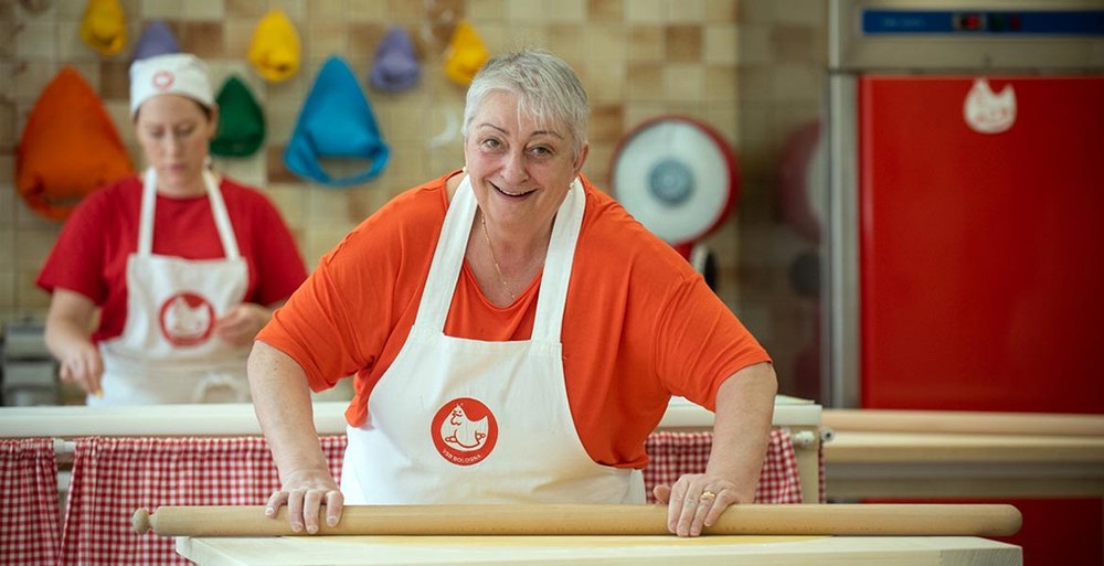 A white-haired woman in an orange top and a white school apron stands behind a long wooden board with both hands resting on a mattarello, looking up and smiling; a second woman in the school's red shirt works at a board behind her, and rows of coloured chef's hats hang on the tiled wall.