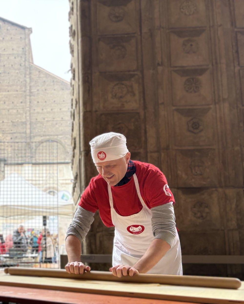 A man in the school's red shirt, white apron and white cap leans over a long wooden board set up outdoors, both hands pushing a mattarello along a sheet of pasta; behind him rises the carved stone front of a Bolognese building, with a crowd watching from the far side.