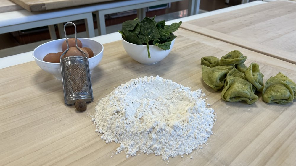 A pale wooden bench with a hand grater, a bowl of brown eggs, a heap of white flour, a white bowl of spinach leaves, and a scatter of green stuffed pasta parcels along the right-hand edge.