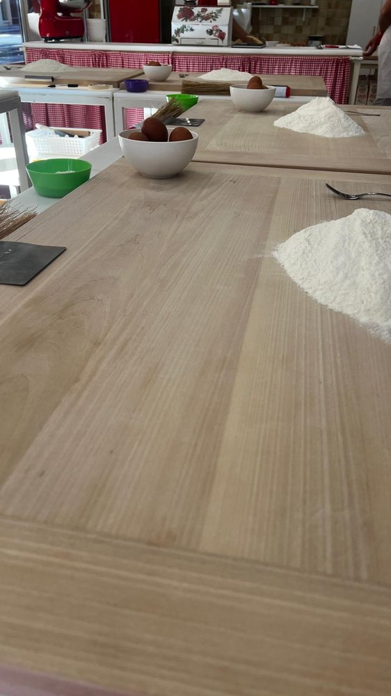 Looking down a long pale wooden board, a heap of white flour on it and a white bowl of brown eggs beyond, with more boards set the same way further down the room and a red-and-white skirted counter at the back.