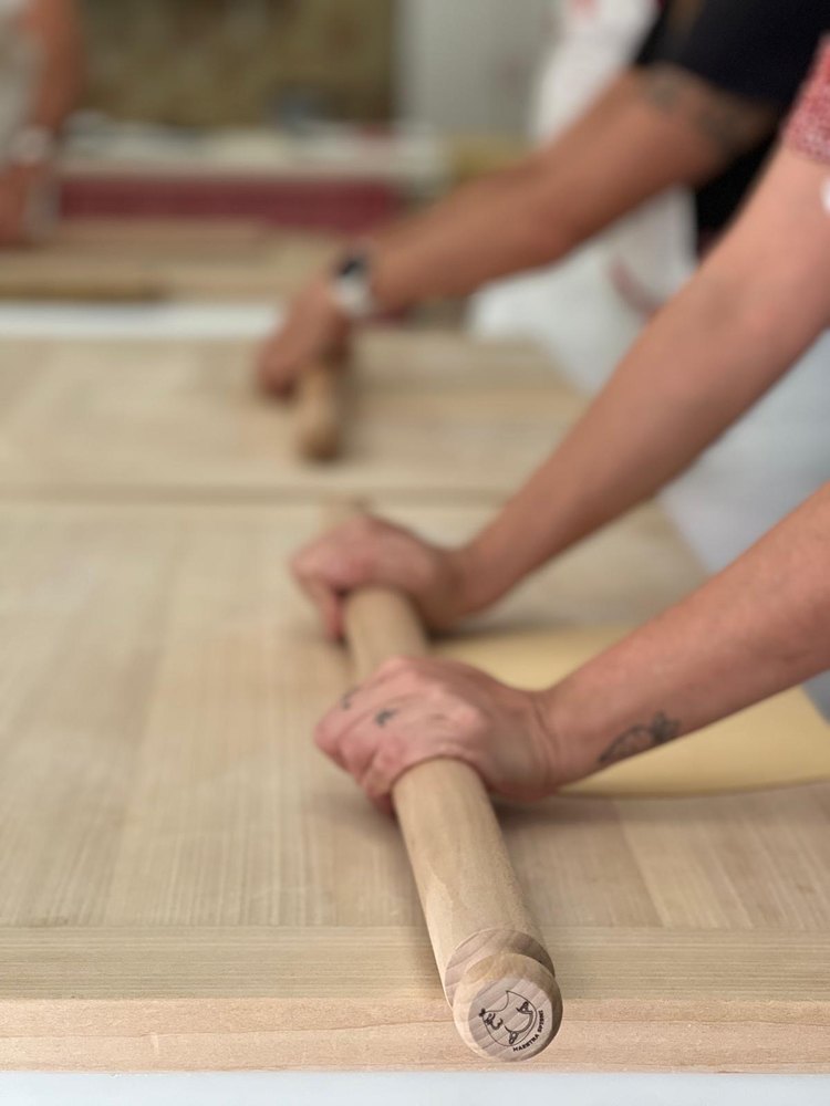 Close on two hands pressing a long wooden mattarello across a yellow sheet of pasta on a wooden board, the school's mark stamped into the end of the pin, and a second pair of hands rolling at the next board behind, out of focus.