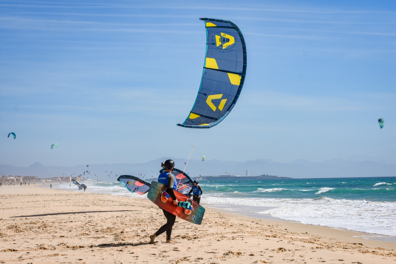 A kitesurfer in a helmet walks down the sand at Tarifa with a board under one arm, their kite already flying overhead, a dozen more kites in the sky behind them and a low headland at the end of the beach.