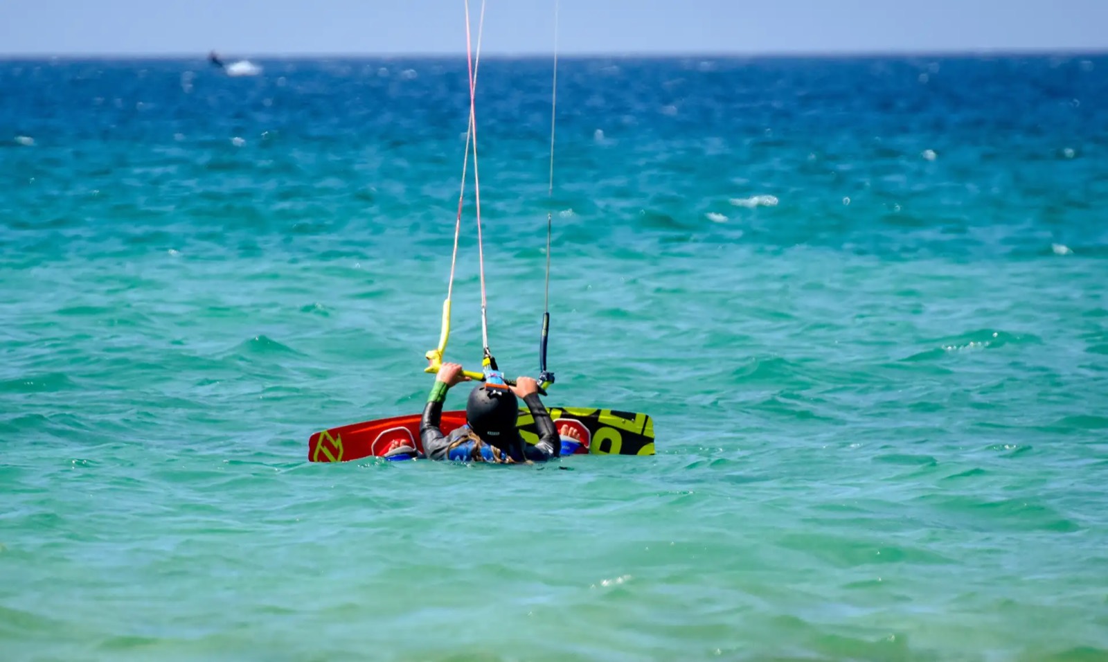 A student in a helmet sits low in shallow turquoise water with a kiteboard across their body, control lines running up out of the frame to the kite.