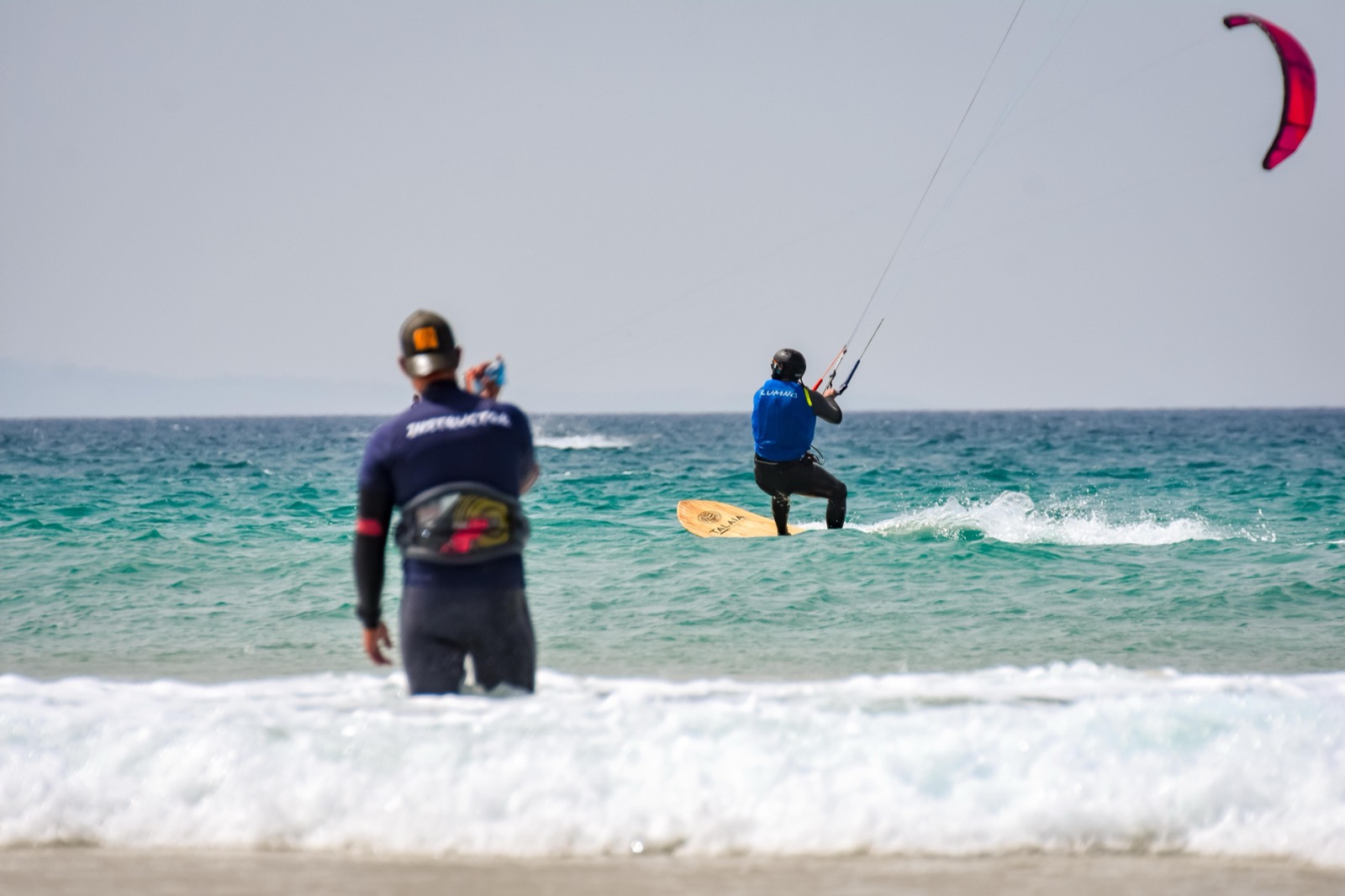 An instructor in a wetsuit lettered INSTRUCTOR across the back stands in the shorebreak watching a helmeted student ride past on a directional board, a kite in the air behind them.