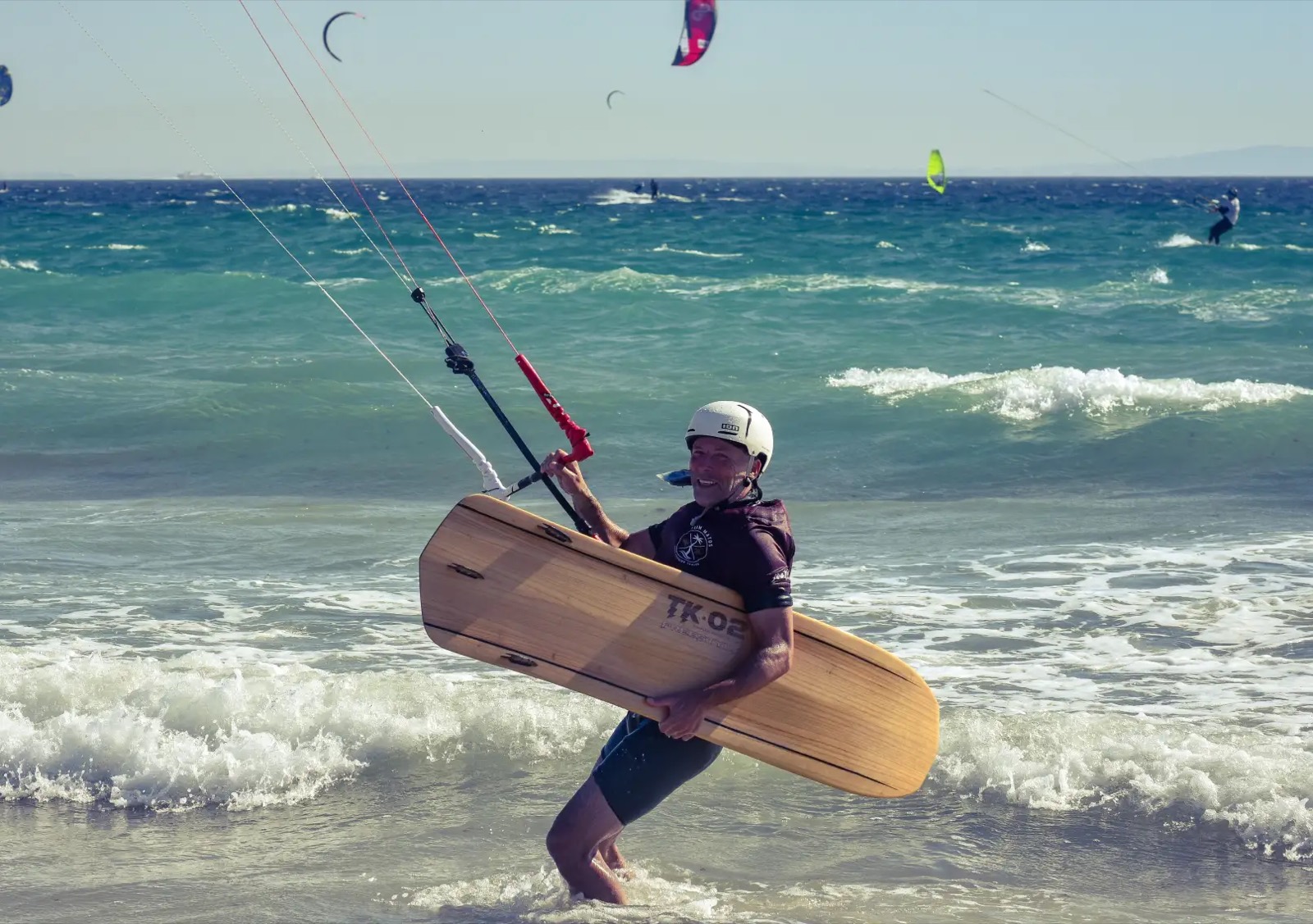 A kitesurfer in a white helmet wades in through the shorebreak carrying a wooden directional board marked TK-02, control bar still in hand, other kites behind.