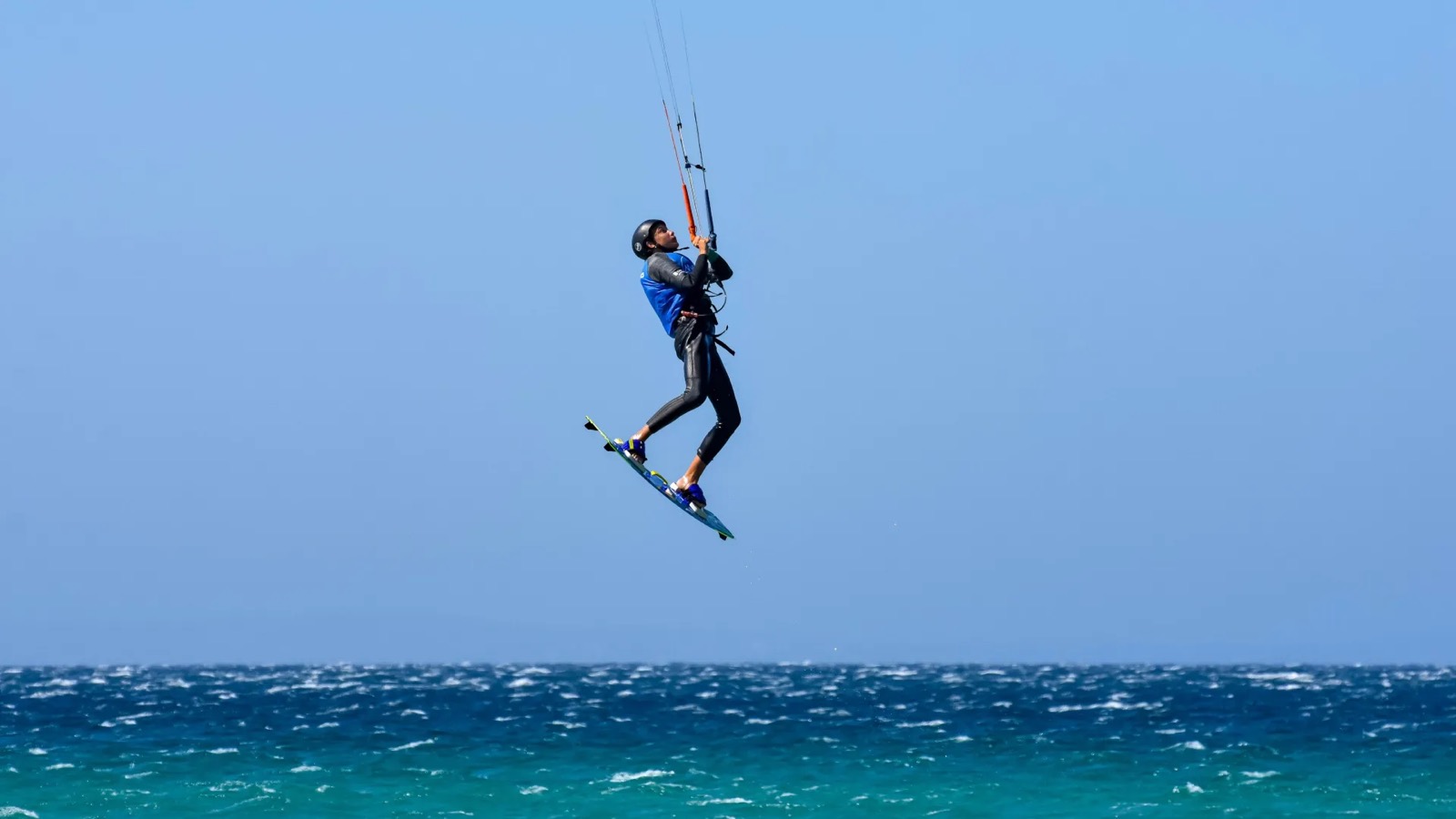 A kitesurfer airborne against a clear sky above choppy blue sea, board still on their feet, head turned up towards the kite.