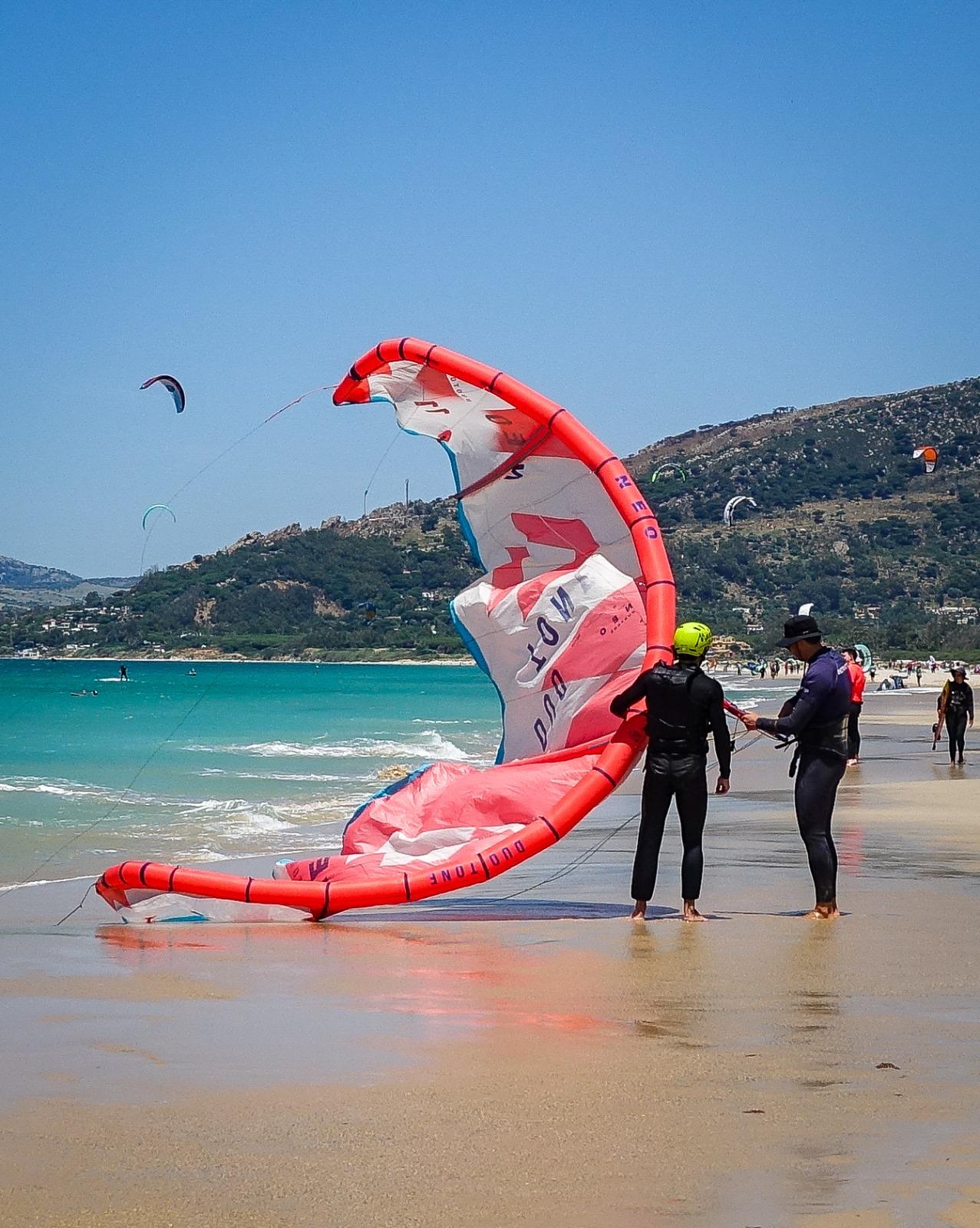 A large red and white kite lies across the wet sand with its lines run out, while an instructor in a wide-brimmed hat holds the bar and a student in a yellow-green helmet stands beside him watching, the hills behind Tarifa and more kites in the sky.