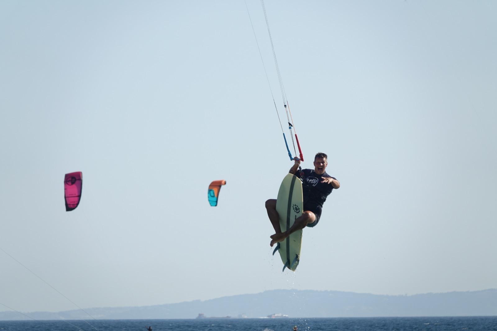 A kitesurfer in the air above the sea, knees drawn up, holding the bar with one hand while a strapless surfboard stays pressed under his bare feet. Two other kites fly further off and a long low coast lies on the horizon.