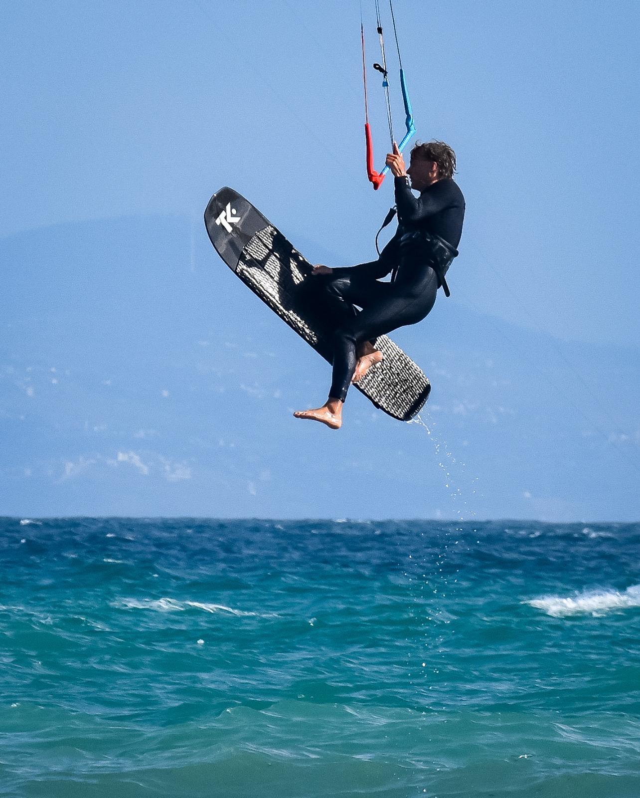A kitesurfer in a black wetsuit high above the sea, knees tucked and the board held flat beneath them, lines running up to a kite out of frame.