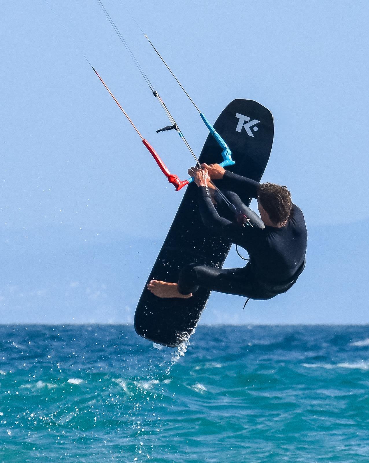 The same rider at the top of a jump, one hand on the control bar and the other reaching down to grab the edge of the board, which is held vertical behind him.