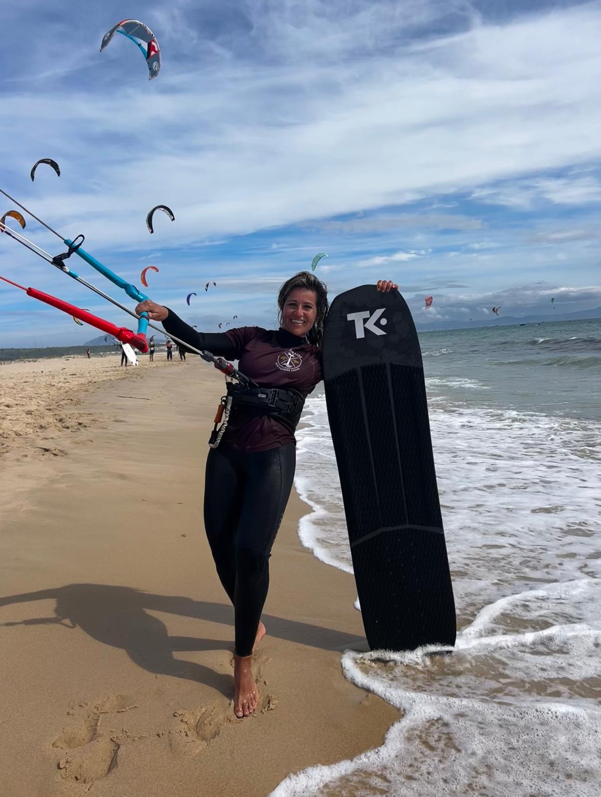 A kitesurfer walks up the wet sand smiling, control bar raised in one hand with the lines running out of frame, a dark board stood upright beside her, a dozen kites in the sky behind.
