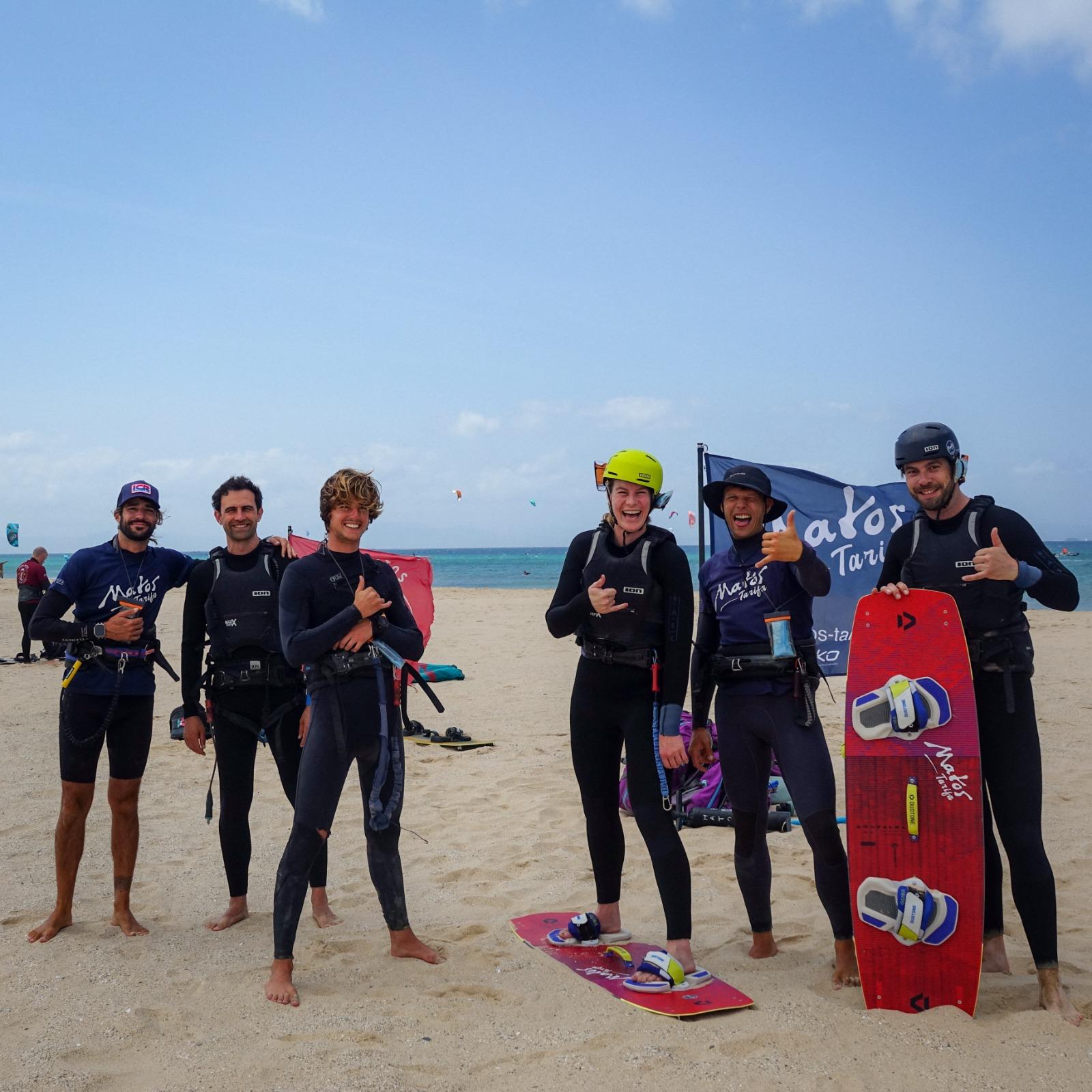 Six people in wetsuits stand on the sand in front of a Matos Tarifa flag, several making a shaka sign, two boards on the ground and a third held upright, kites in the sky behind them.