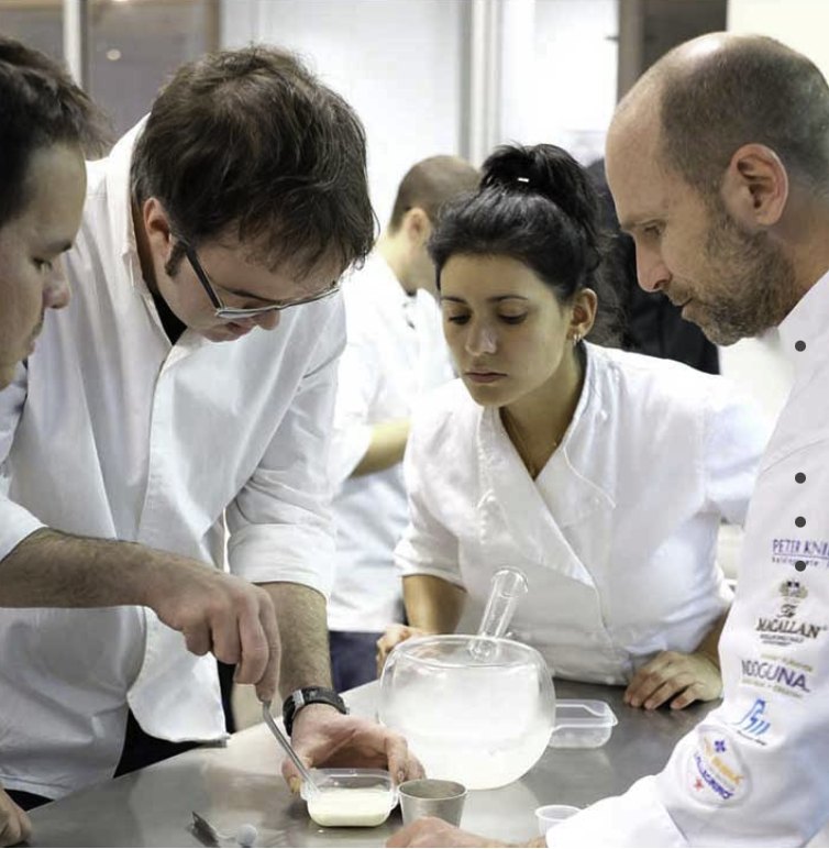 Several cooks in white jackets lean in close together over a steel bench; one in glasses works a small spoon into a metal cup beside a large glass jug of white liquid, a dark-haired woman watching from beside him and another man looking down at the work.
