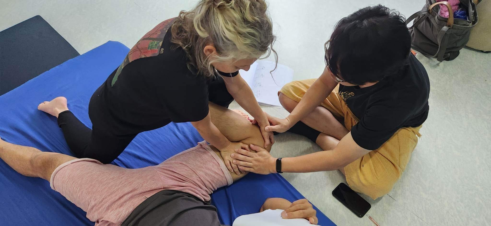 Seen from above: a student kneeling over a person lying on a blue floor mat presses into their thigh with both hands, while a second pair of hands reaches in from the right and rests on top of hers, guiding the pressure. An open notebook lies on the floor beside them.
