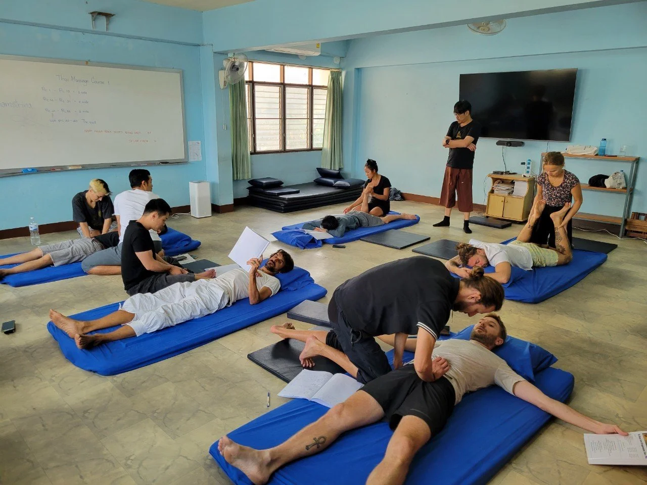 A classroom with blue floor mats laid out in rows, six pairs of students working on each other, an instructor standing at the back, and a whiteboard reading “Thai Massage Course 1”.