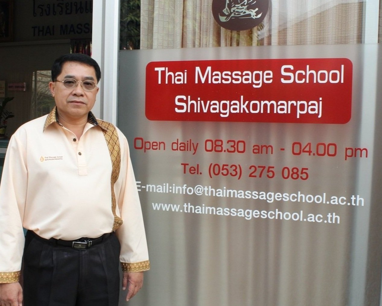 A man in a cream Thai shirt with a gold sash over one shoulder stands beside the school's sign at its entrance. The sign reads Thai Massage School Shivagakomarpaj, open daily 08.30 am to 04.00 pm, with a telephone number, an email address and the school's website.
